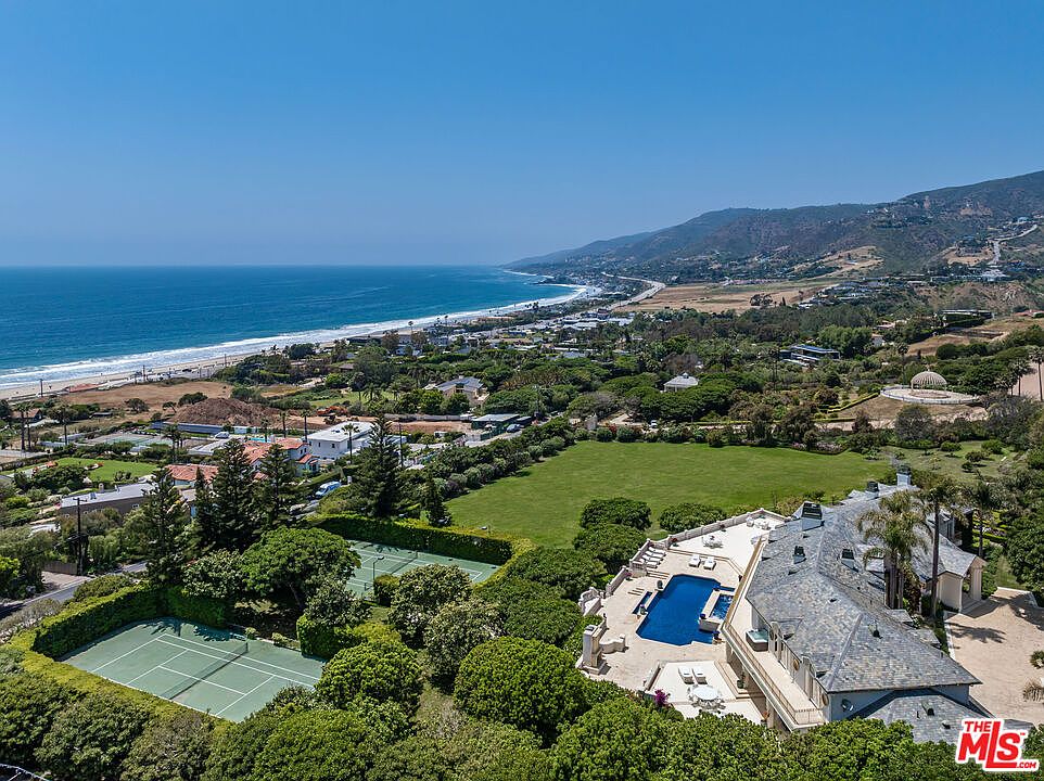 This stunning aerial perspective captures a sprawling luxury estate overlooking the Pacific coastline. The property features a large private swimming pool, a tennis court, and expansive manicured lawns, all set against a backdrop of rolling hills and the vast ocean. The cinematic high-angle shot emphasizes the grandeur of the residence and its prime coastal location.