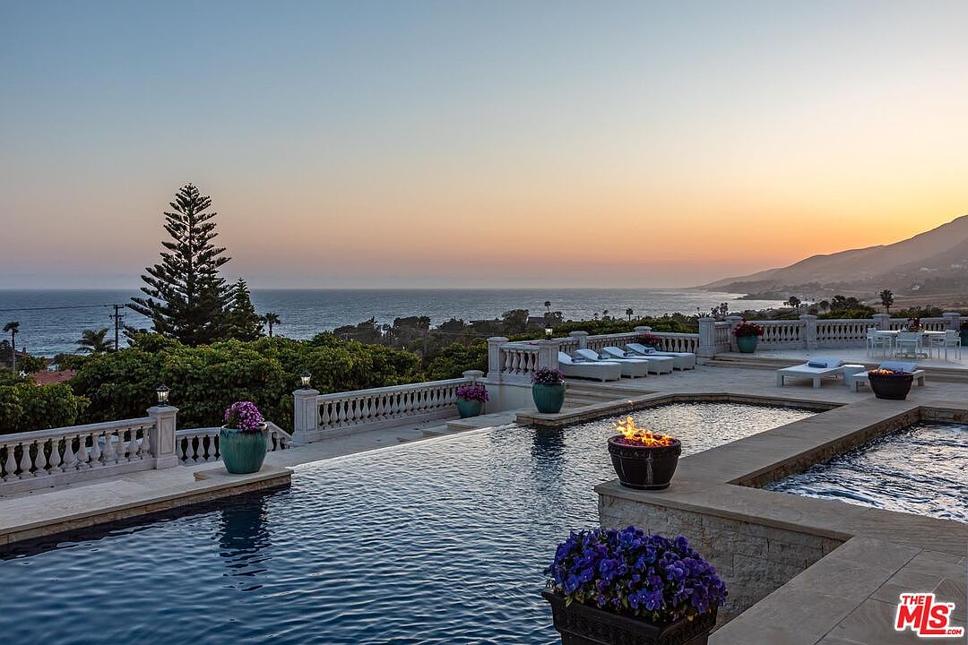 This stunning infinity-edge pool overlooks a breathtaking coastal sunset, creating a seamless visual connection between the water and the horizon. The expansive stone terrace features elegant balustrades, multiple lounge chairs, and decorative fire bowls that add a touch of luxury to the outdoor living space. The scene is framed by vibrant purple flowers and lush greenery, emphasizing a sophisticated and serene atmosphere perfect for high-end coastal living.