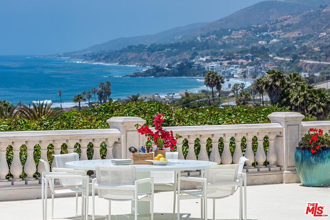 This elegant outdoor terrace features a classic white stone balustrade and a matching dining set, perfectly positioned to capture breathtaking panoramic views of the coastline and ocean. The scene is accented by vibrant bougainvillea flowers and a large, decorative ceramic planter, creating a sophisticated and serene atmosphere. The perspective is eye-level, emphasizing the seamless transition between the luxurious living space and the expansive natural landscape beyond.