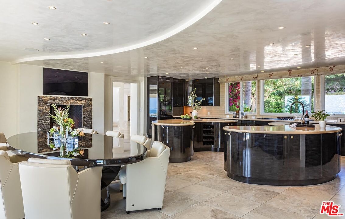 This expansive, open-concept kitchen features a unique, high-gloss black curved island and matching cabinetry that contrasts elegantly with the light-toned tile flooring. A large, oval dining table sits adjacent to a fireplace with a mounted television, creating a seamless transition between cooking and dining areas. The room is illuminated by recessed lighting set into a sophisticated, curved ceiling design, offering a luxurious and modern aesthetic.