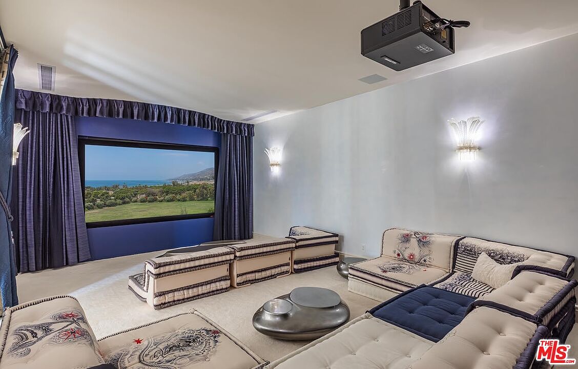 This spacious home theater features a large projection screen framed by deep blue curtains, creating a cinematic atmosphere. The room is furnished with modular, low-profile seating upholstered in patterned fabric, centered around a unique, metallic, multi-tiered coffee table. A ceiling-mounted projector and elegant wall sconces provide functional lighting, while the overall aesthetic blends modern comfort with a touch of luxury.