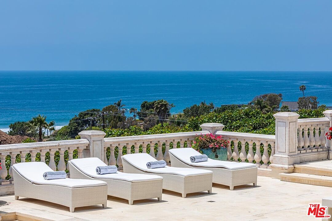 This expansive stone terrace features four elegant white lounge chairs arranged in a row, each topped with a neatly folded towel, overlooking a breathtaking panoramic view of the ocean. The space is enclosed by a classic stone balustrade, accented by a vibrant potted plant, creating a luxurious and serene atmosphere perfect for relaxation. The perspective is eye-level, capturing the seamless transition from the refined patio architecture to the vast, tranquil horizon.