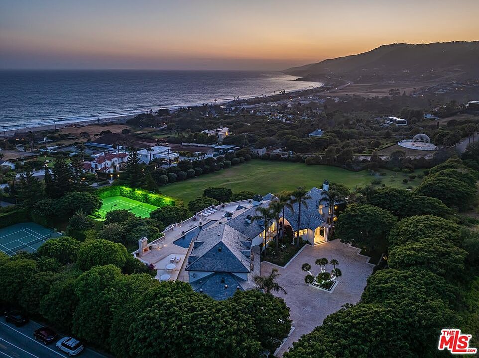 This stunning aerial view captures a sprawling luxury estate nestled along the coastline at sunset. The property features a large slate-roofed mansion, a private tennis court, a swimming pool, and expansive manicured lawns, all surrounded by lush greenery. The cinematic perspective highlights the seamless integration of high-end residential living with the breathtaking natural beauty of the oceanfront landscape.