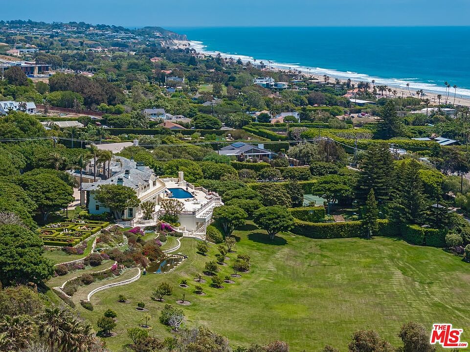 This stunning aerial view captures a sprawling coastal estate featuring a grand multi-story residence with a private swimming pool and meticulously landscaped gardens. The property transitions from lush, manicured lawns and structured flower beds into a dense canopy of trees, all set against the backdrop of a vast, sparkling ocean coastline. The perspective emphasizes the expansive scale of the grounds and the prime, secluded location of this luxury home.