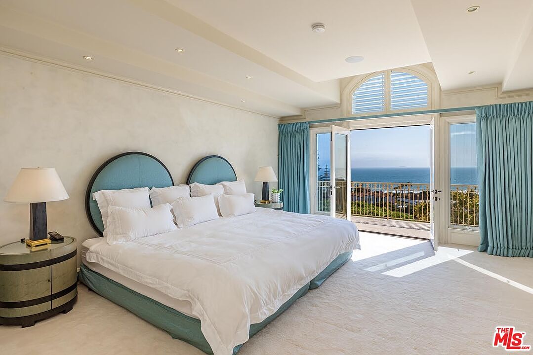 This luxurious primary bedroom features a large, plush bed with a distinctive dual-arched, light blue upholstered headboard as the focal point. The room is bathed in natural light from expansive glass doors that open onto a private balcony, offering a stunning, unobstructed view of the ocean. The space is decorated in a serene, coastal-inspired palette with soft cream carpeting and elegant blue curtains, creating a tranquil and sophisticated atmosphere.