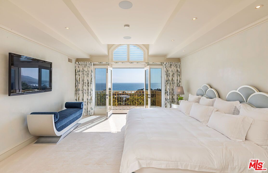 This luxurious primary bedroom features a spacious layout with a large, plush bed adorned with white linens and a unique scalloped headboard. A sleek, modern blue velvet chaise lounge sits beneath a wall-mounted television, while floor-to-ceiling glass doors open onto a private balcony with stunning ocean views. The room is bathed in natural light, creating an airy, serene atmosphere that emphasizes high-end coastal living.