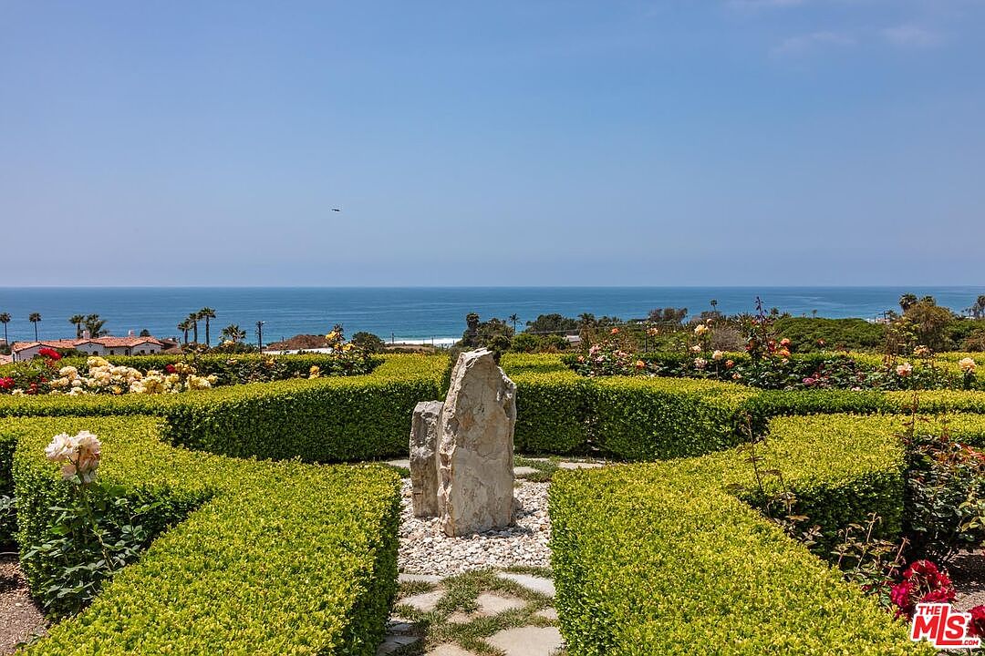 This beautifully manicured garden features a formal geometric hedge design surrounding a central, weathered stone sculpture. The foreground showcases vibrant rose bushes, while the background offers a breathtaking, unobstructed panoramic view of the Pacific Ocean under a clear blue sky. The perspective is eye-level, capturing the serene transition from the structured landscape to the vast horizon.