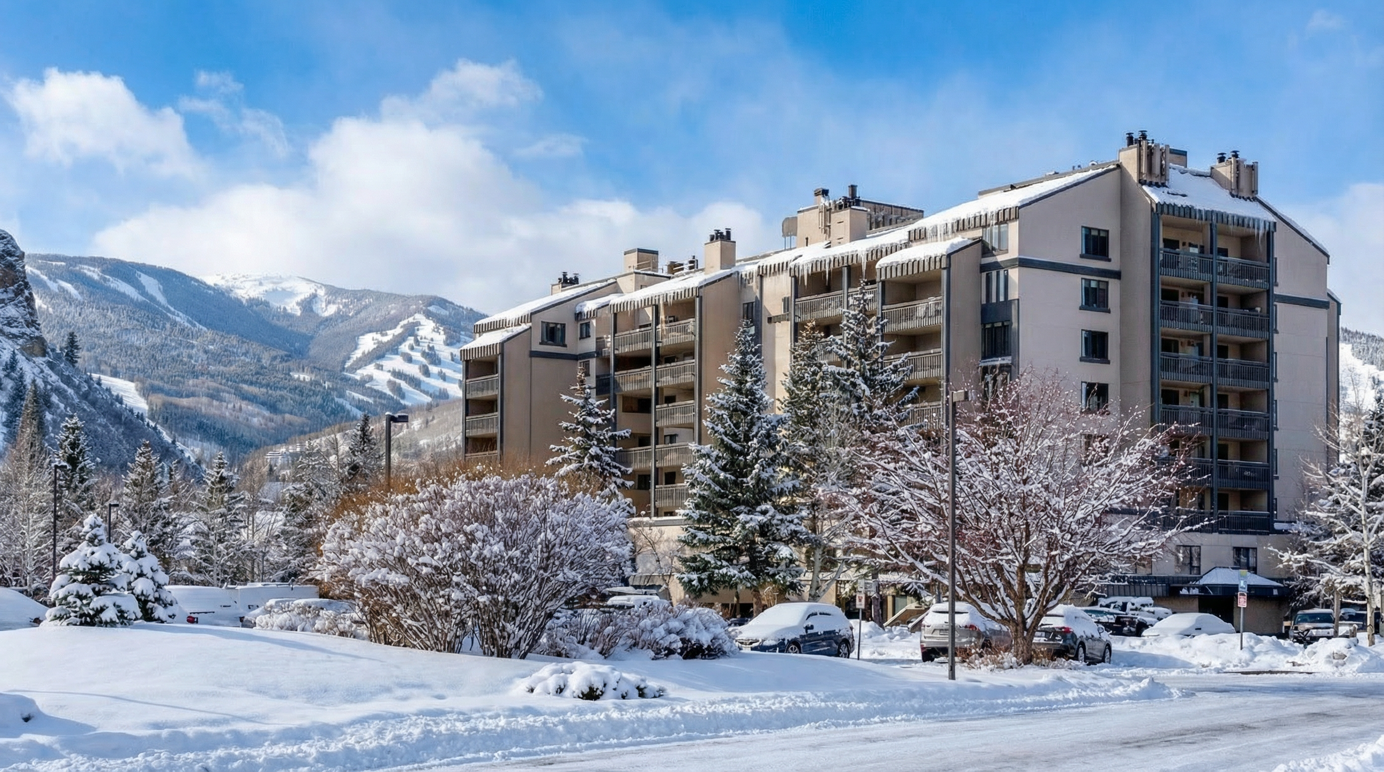 This image presents a side-front perspective of a multi-story condominium or apartment building situated in a snowy, mountainous landscape. The architecture features multiple tiers of private balconies and a neutral-toned facade, complemented by mature evergreen trees in the foreground. The setting provides a picturesque alpine feel, making it an appealing residence for ski enthusiasts and those seeking a scenic mountain escape.