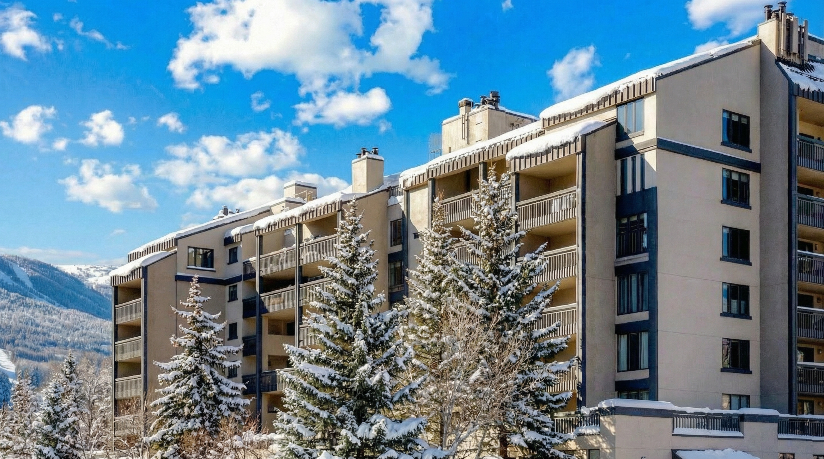 This image shows a multi-story mountain condominium complex during winter with snow-covered roofs and surrounding pine trees. The building features a tiered architectural design with numerous balconies and large windows that offer scenic views of the nearby snowy slope. The exterior is finished in a neutral tone with dark framing elements, conveying a cozy, alpine lodge aesthetic ideal for a seasonal retreat.