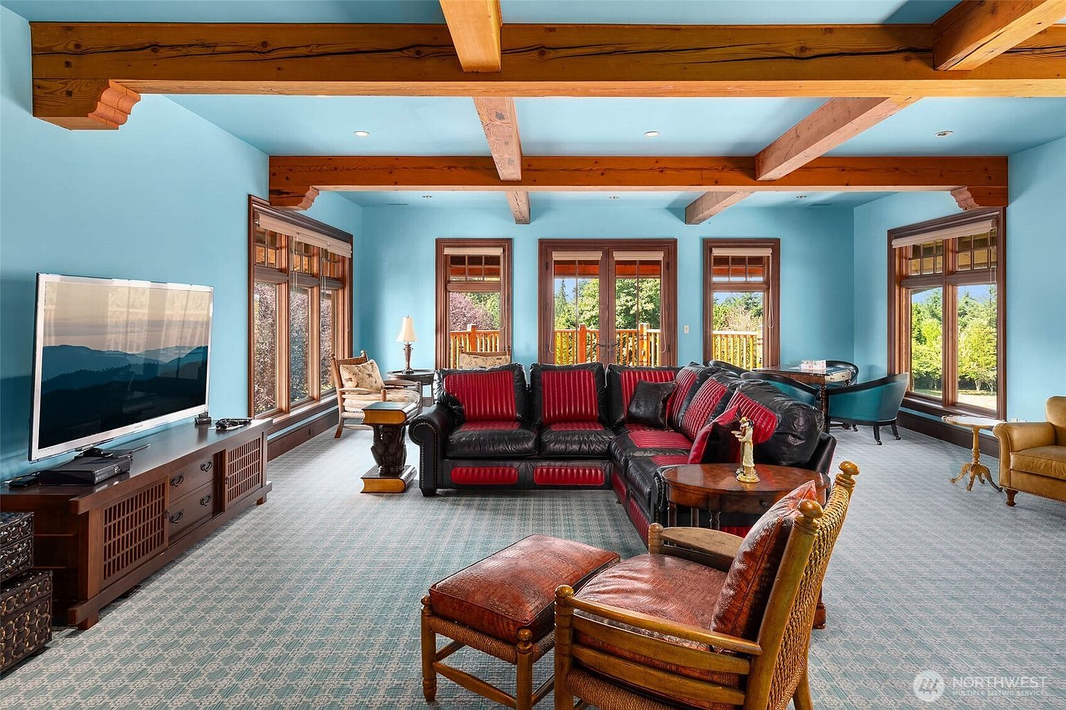 This spacious living room features a distinctive blue wall color contrasted by exposed wooden ceiling beams and dark wood window trim. The room is anchored by a large, multi-textured sectional sofa in black and red, complemented by a patterned carpet and a leather armchair with a matching ottoman. Large windows provide ample natural light and views of the surrounding greenery, creating a cozy yet expansive atmosphere.