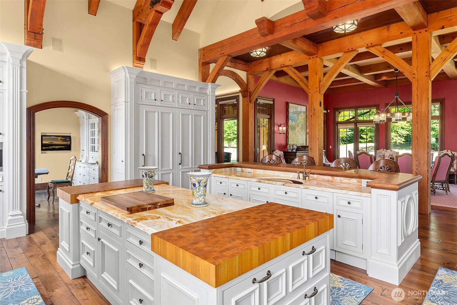 This spacious kitchen features a large central island with a light-toned stone countertop and a thick wood-block prep area, complemented by white cabinetry and exposed timber ceiling beams. The open-concept design flows into a dining area with deep red walls and a view of the outdoors through large windows. The perspective captures the intricate craftsmanship of the woodwork and the warm, inviting atmosphere of the home.