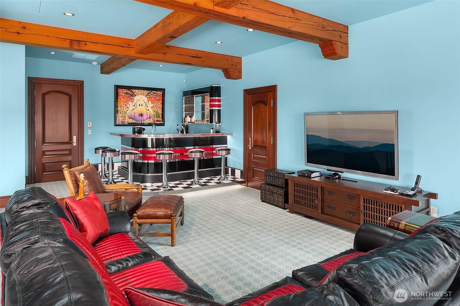 This spacious basement recreation room features a retro-themed bar area with a black and red color scheme, checkered flooring, and bar stools. The room is anchored by a large, plush leather sectional sofa facing a television mounted on a wooden media console, all set beneath exposed wooden ceiling beams. The light blue walls and patterned carpeting create a casual, entertainment-focused atmosphere perfect for hosting guests.