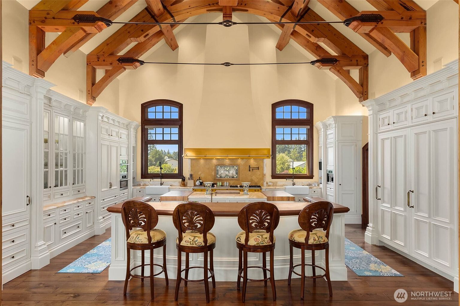 This expansive, high-ceilinged kitchen features a symmetrical layout with white cabinetry, a large central island with bar seating, and exposed timber trusses overhead. Two large windows flank a central cooking area, which is accented by a prominent metallic hood and a decorative tile backsplash. The room is anchored by dark wood flooring and a long wooden countertop on the island, creating a sophisticated and airy atmosphere.