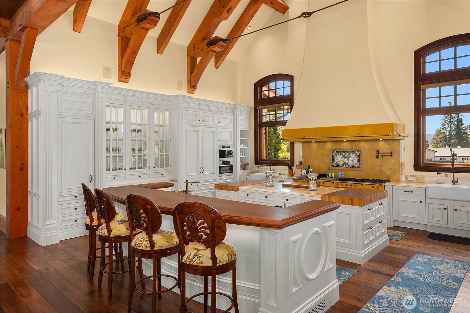 This spacious, high-ceilinged kitchen features a large central island with a butcher-block countertop and a matching breakfast bar with three wooden stools. The room is characterized by white cabinetry, a prominent yellow-toned range hood, and exposed wooden ceiling beams that add a rustic, architectural flair. Large windows allow natural light to fill the space, highlighting the warm wood flooring and the intricate, patterned area rug.