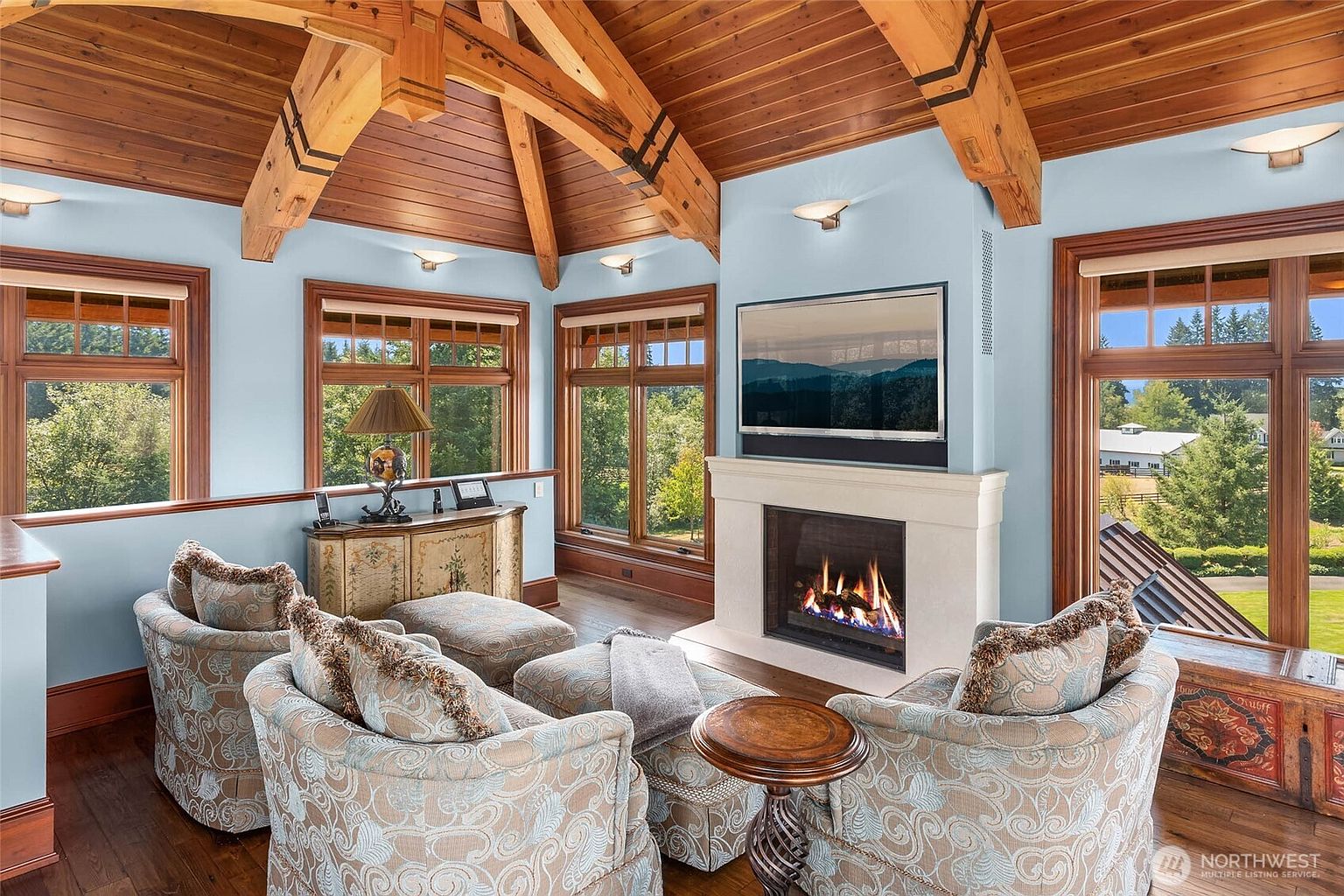 This cozy living room features a vaulted ceiling with exposed timber beams and warm wood paneling. A central fireplace with a light-colored stone surround serves as the focal point, flanked by large windows that provide abundant natural light and views of the surrounding landscape. The space is furnished with comfortable patterned armchairs and ottomans, creating an inviting atmosphere for relaxation.