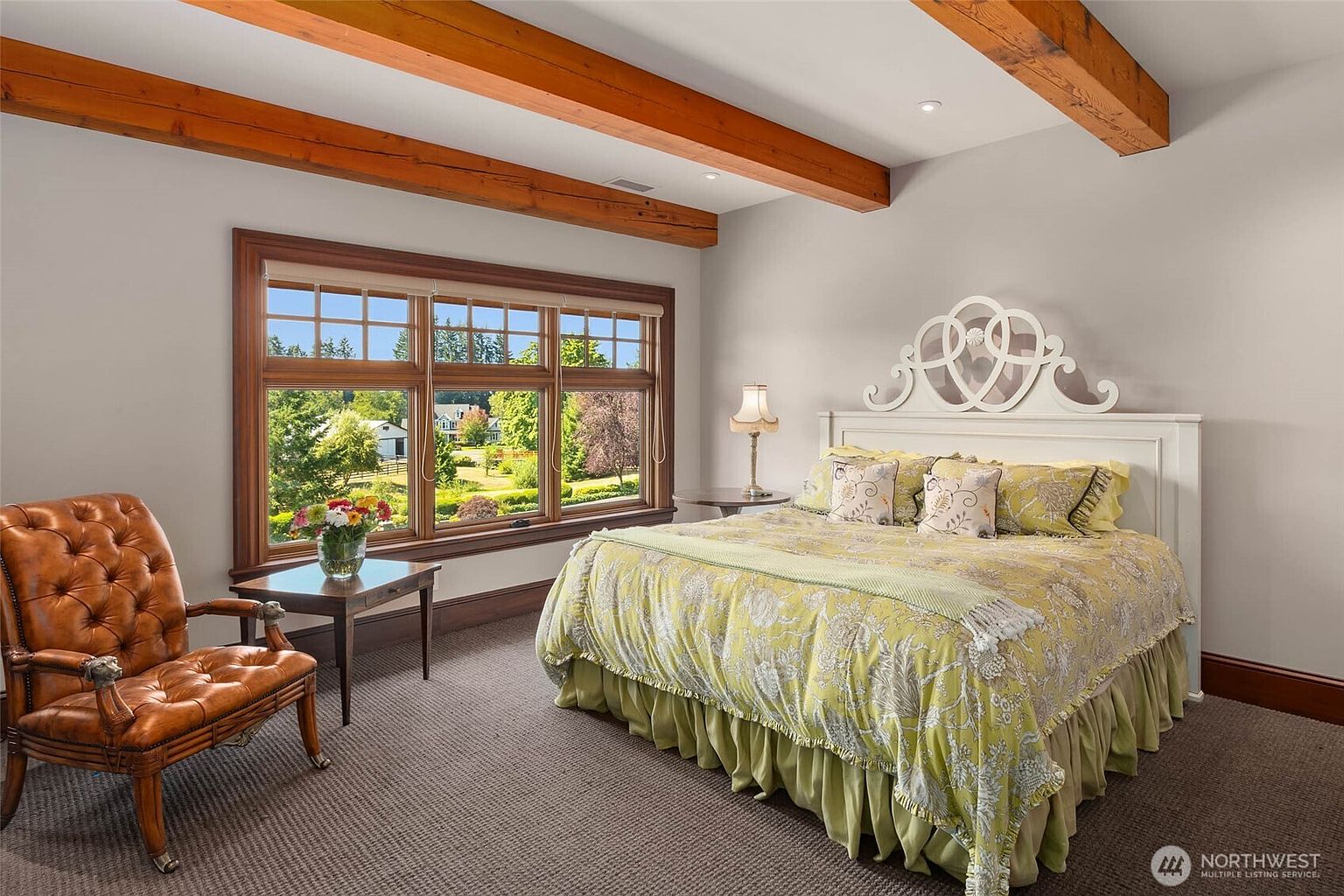This spacious bedroom features a large bed with a decorative white headboard and a yellow patterned quilt, positioned near a set of windows that offer a scenic view of the outdoors. A tufted leather armchair sits in the corner next to a small side table, while exposed wooden ceiling beams add a rustic architectural element to the room. The space is finished with neutral wall tones and wall-to-wall carpeting, creating a warm and inviting atmosphere.