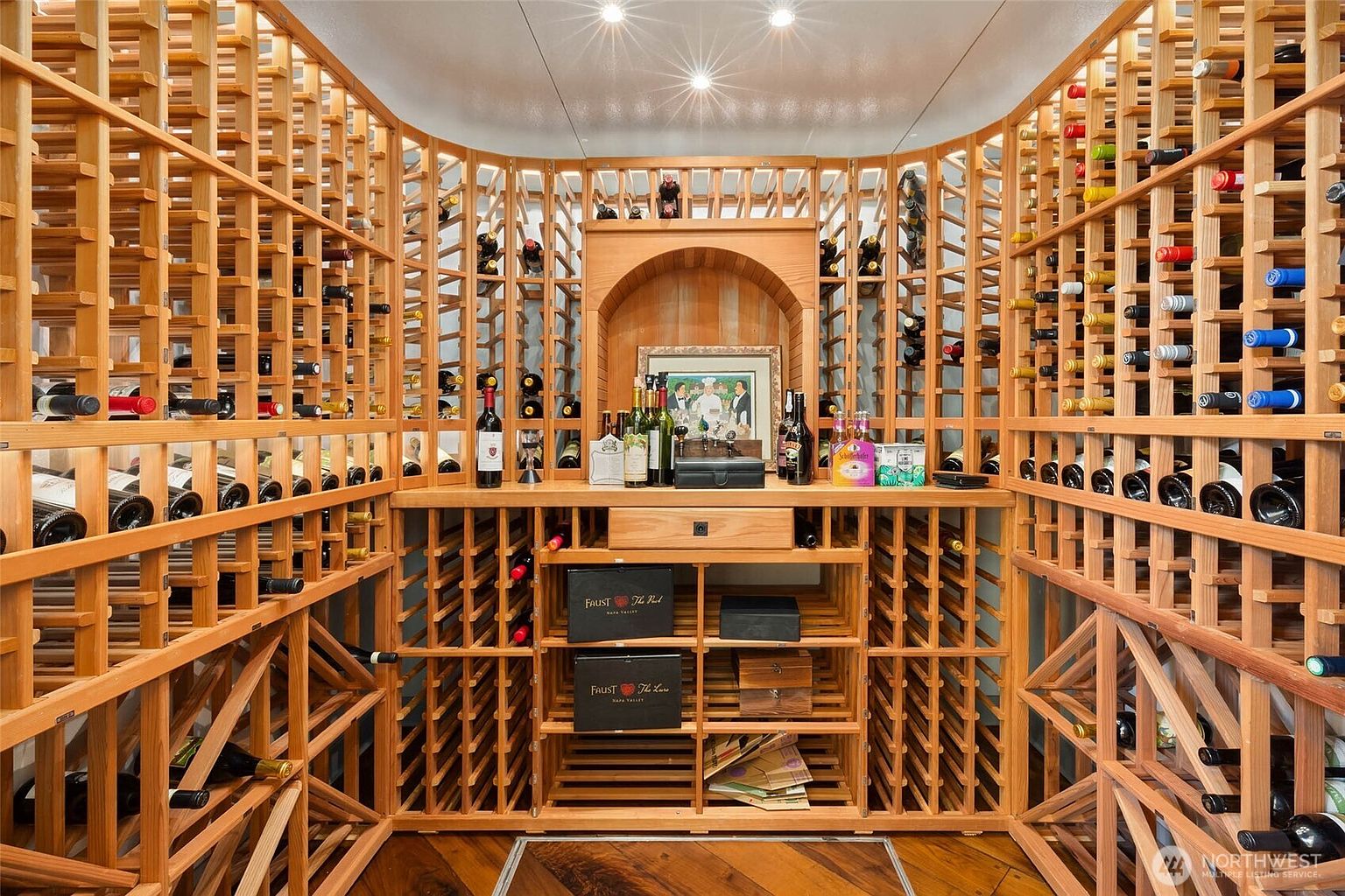This custom-built wine cellar features extensive light wood racking that spans the walls, providing ample storage for a large collection. A central counter area includes a small drawer and a framed picture, creating a focal point within the symmetrical layout. The space is illuminated by recessed ceiling lights, highlighting the organized arrangement of bottles and wooden crates.