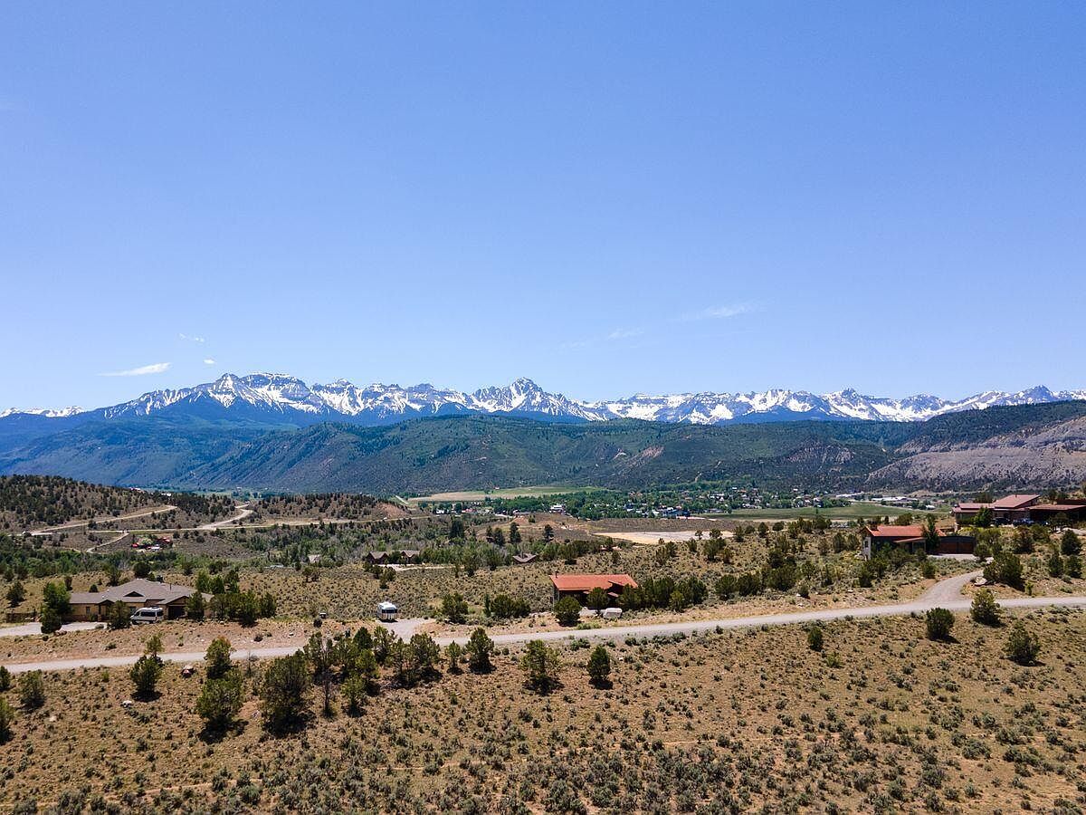 This aerial view showcases a sprawling landscape with scattered homes and winding roads. In the background, a majestic mountain range with snow-capped peaks dominates the horizon under a clear blue sky. The foreground features arid terrain with sparse vegetation, creating a contrast between the developed areas and the natural environment.