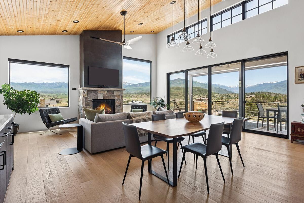 This is an interior shot of a modern living room and dining area featuring an open floor plan. The room boasts high ceilings with wood paneling, large windows offering scenic mountain views, and a stone fireplace with a mounted television. The dining area includes a wooden table with black chairs, and the living room has a comfortable gray sofa and a stylish black chair.