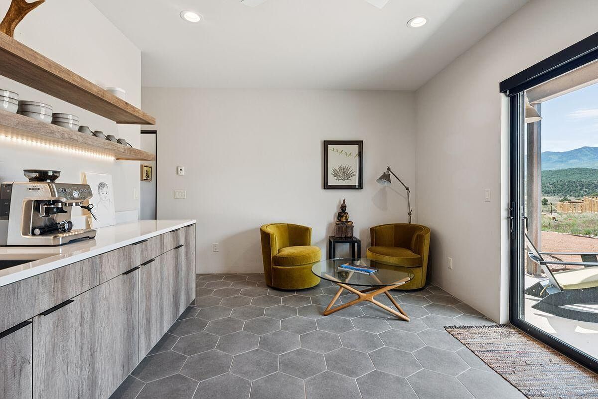 This interior shot showcases a cozy living area adjacent to a kitchen space, featuring stylish gray hexagonal floor tiles and a pair of mustard-colored armchairs flanking a glass coffee table. A sliding glass door opens to an outdoor patio with mountain views, enhancing the room's appeal with natural light and a connection to the outdoors. The room is decorated with a framed print and a small statue on a side table, adding a touch of personality.