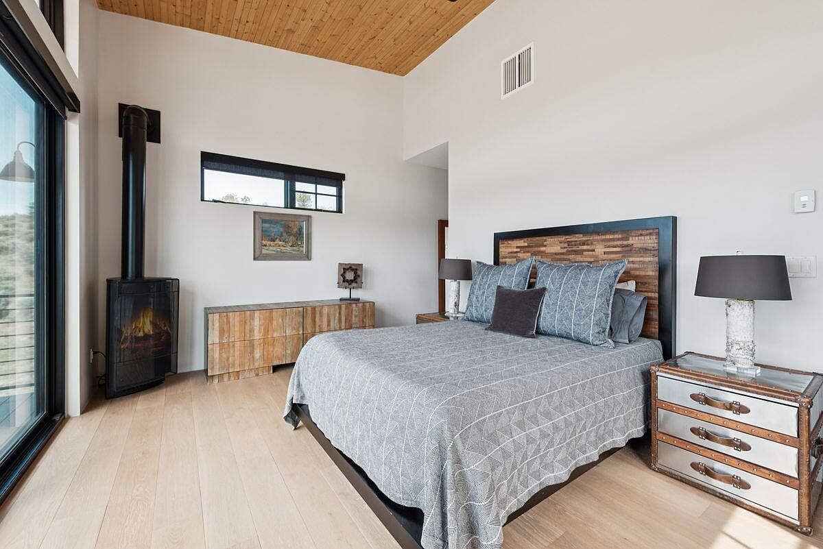 This is a well-lit primary bedroom featuring a modern rustic design. The room includes a bed with a wooden headboard and gray patterned bedding, a fireplace, and wooden flooring. The room also has a large window and a small window, allowing for natural light.
