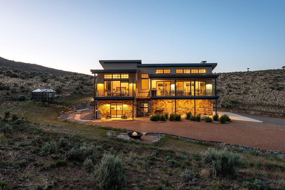 This is a rear exterior view of a modern two-story home built into a hillside. The home features a stone foundation, large windows with balconies on the upper level, and a fire pit in the yard. The surrounding landscape is natural and rugged, with a greenhouse visible in the distance.