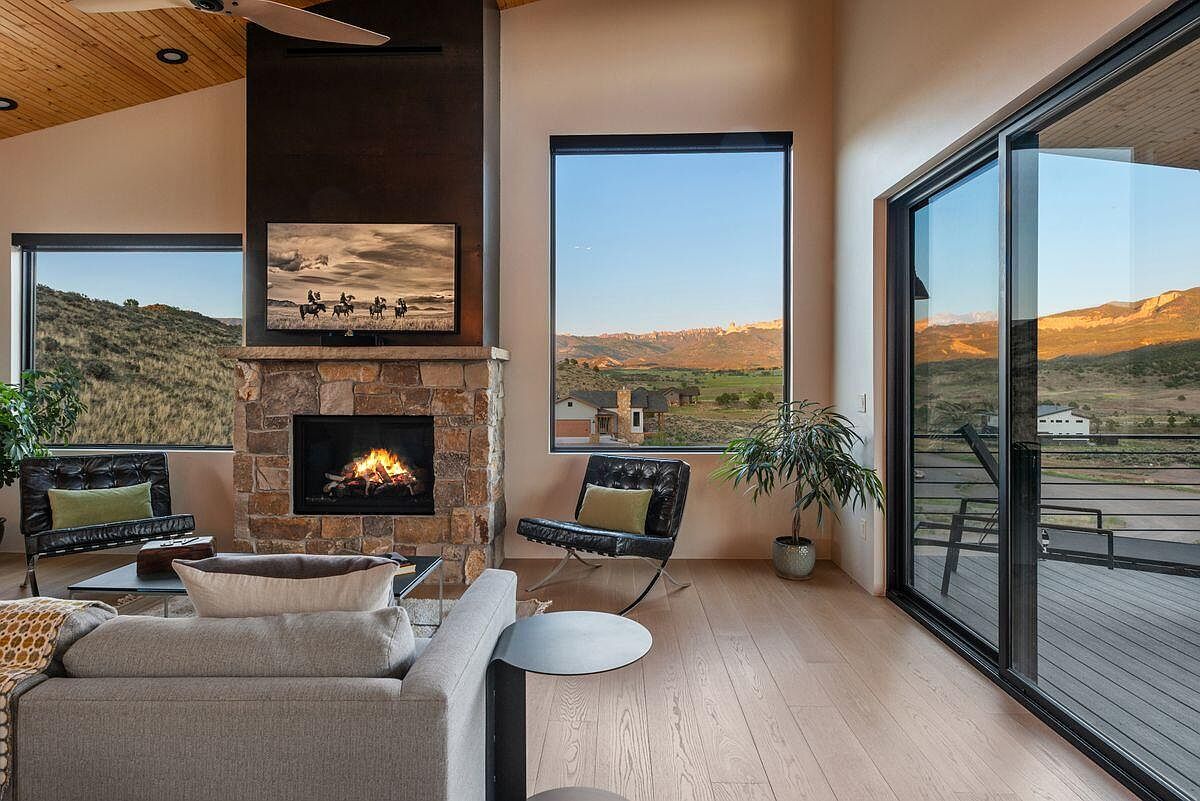 This is a beautifully staged living room featuring a stone fireplace with a mounted television above. The room is well-lit with natural light from large windows offering scenic views of the surrounding landscape. The space is furnished with modern seating and stylish decor, creating a warm and inviting atmosphere.