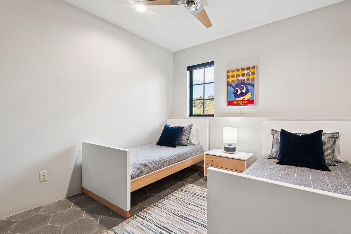 This is a bright and modern guest bedroom featuring two twin beds with white frames and striped bedding. A colorful piece of art hangs above one of the beds, adding a playful touch. The room is well-lit with a window and a bedside lamp, and the gray hexagonal tile flooring and patterned rug add visual interest.