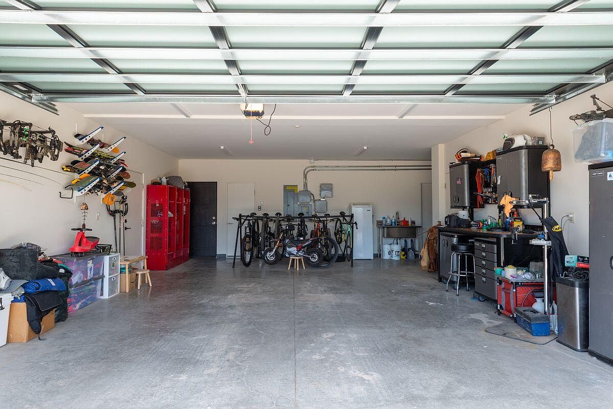 This is a spacious and well-organized garage. The garage features a clear glass panel ceiling, providing ample natural light. The space is equipped with storage solutions, including red lockers, wall-mounted racks for skis and bows, and a workbench area with cabinets and a refrigerator, making it ideal for hobbies and storage.