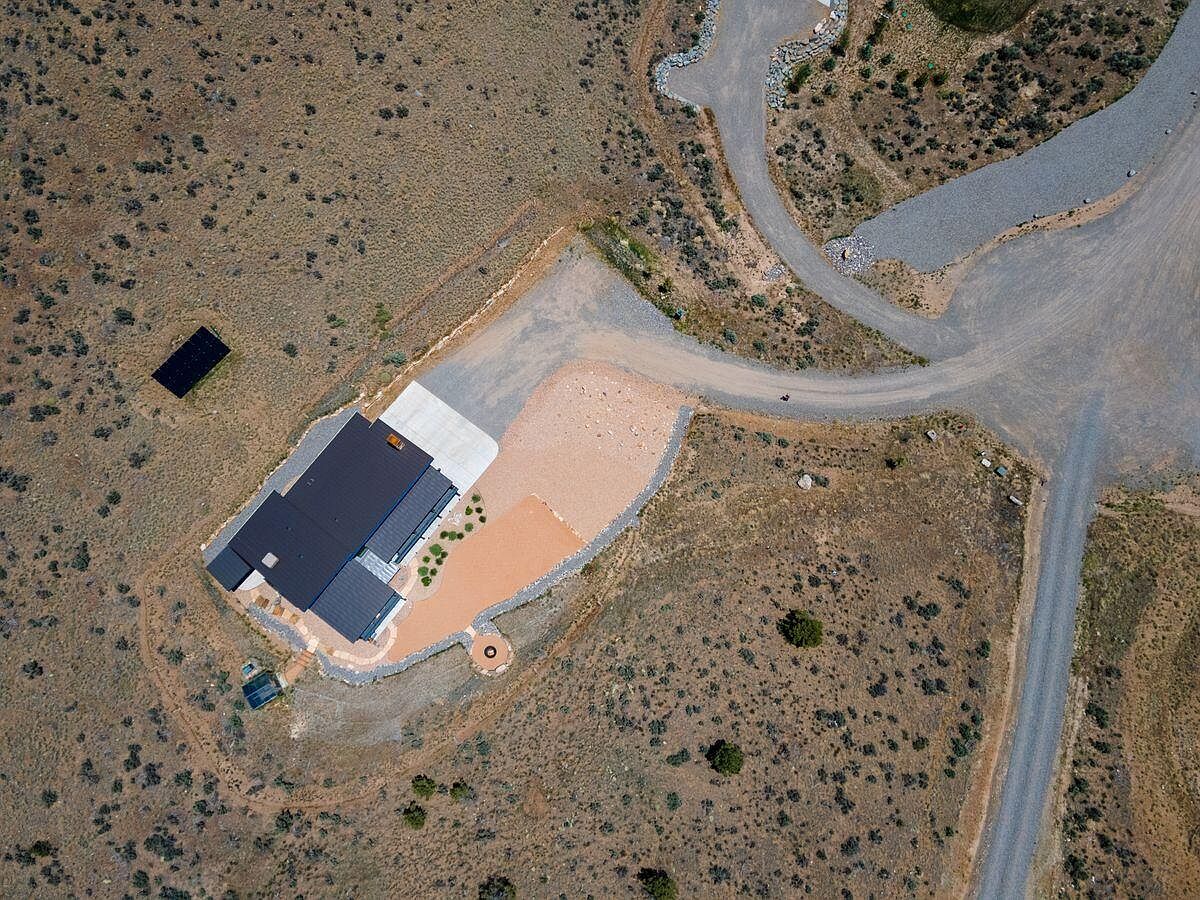 This aerial shot showcases a modern home nestled in a natural landscape. The house features a dark roof, a concrete driveway, and a surrounding gravel area. A winding dirt road leads to the property, and a separate solar panel installation is visible nearby, highlighting the property's integration with its environment.