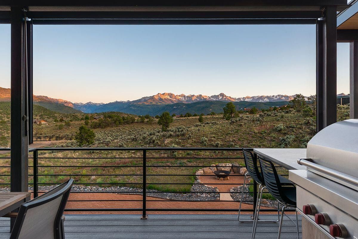 This image showcases a modern patio or deck with a stunning mountain view. The space features a built-in grill, sleek outdoor furniture, and a fire pit area, creating an inviting atmosphere for relaxation and entertainment. The deck is constructed with dark wood and has a black metal railing.
