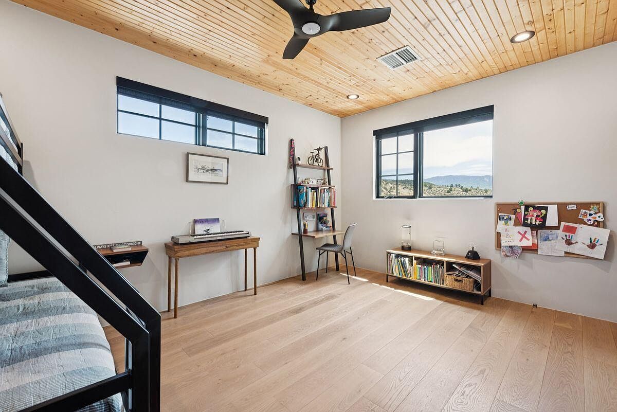 This is a bright and airy bedroom featuring light hardwood floors and a wood-paneled ceiling with a modern ceiling fan. The room includes two windows that provide natural light, a desk area with a ladder bookshelf, and a corkboard with artwork. A bunk bed is partially visible on the left, suggesting a child's or guest room.