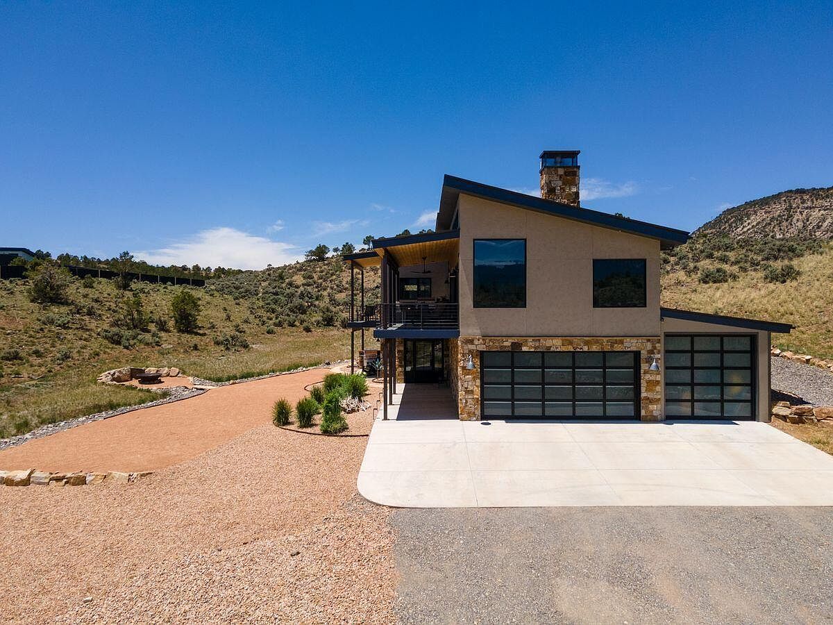 This is an eye-level, slightly elevated front view of a modern two-story home with a two-car garage. The house features a combination of stucco and stone facade, with large windows and a dark-colored roof. The driveway is concrete, and the surrounding landscape is a mix of gravel and natural vegetation, blending seamlessly with the hillside setting.