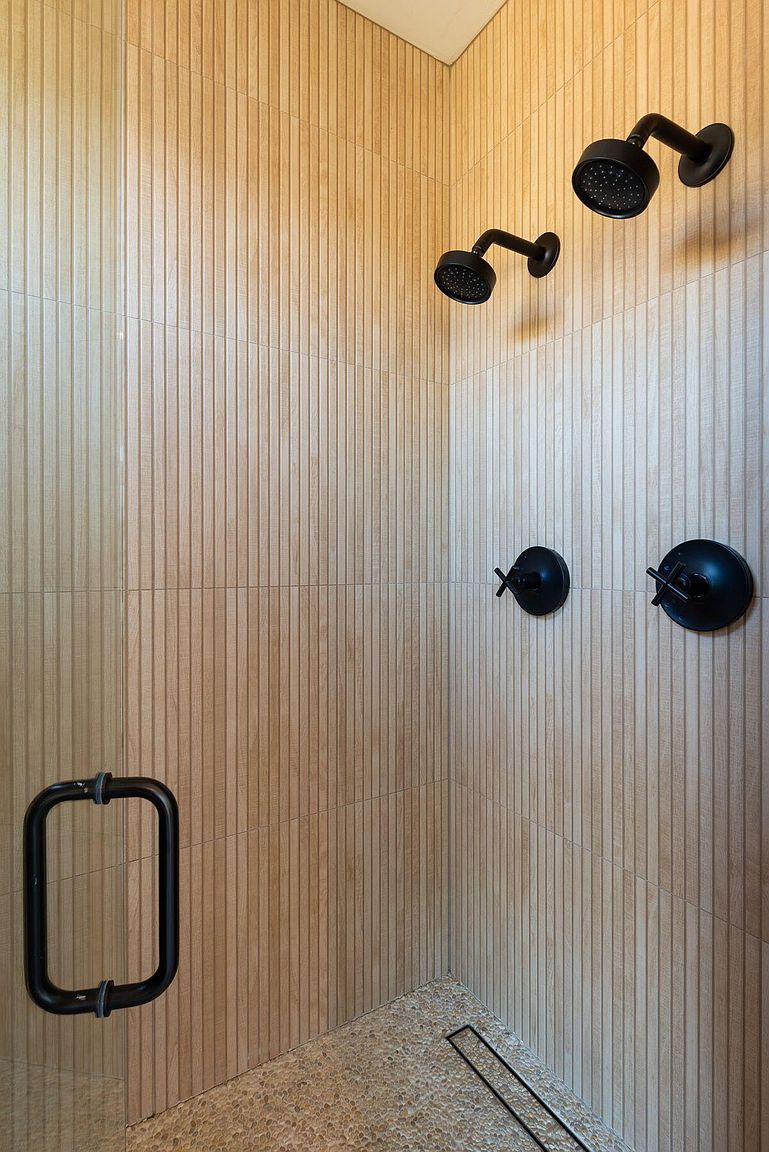 This is a modern primary bathroom featuring a shower with vertically-aligned, light-colored, textured tiles. The shower is equipped with multiple black shower heads and matching black fixtures. A black shower door handle is visible on the left, and the shower floor appears to be made of small pebbles.