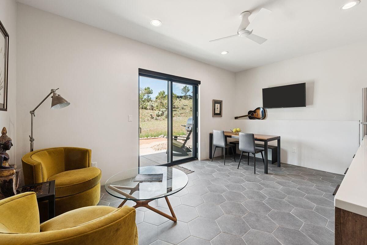This interior shot showcases a modern living room with a focus on natural light and contemporary design. The room features a sliding glass door leading to an outdoor area, a dining table with chairs, and comfortable yellow armchairs around a glass coffee table. The flooring is a stylish gray hexagon tile, and the walls are painted in a neutral tone, creating a bright and inviting atmosphere.