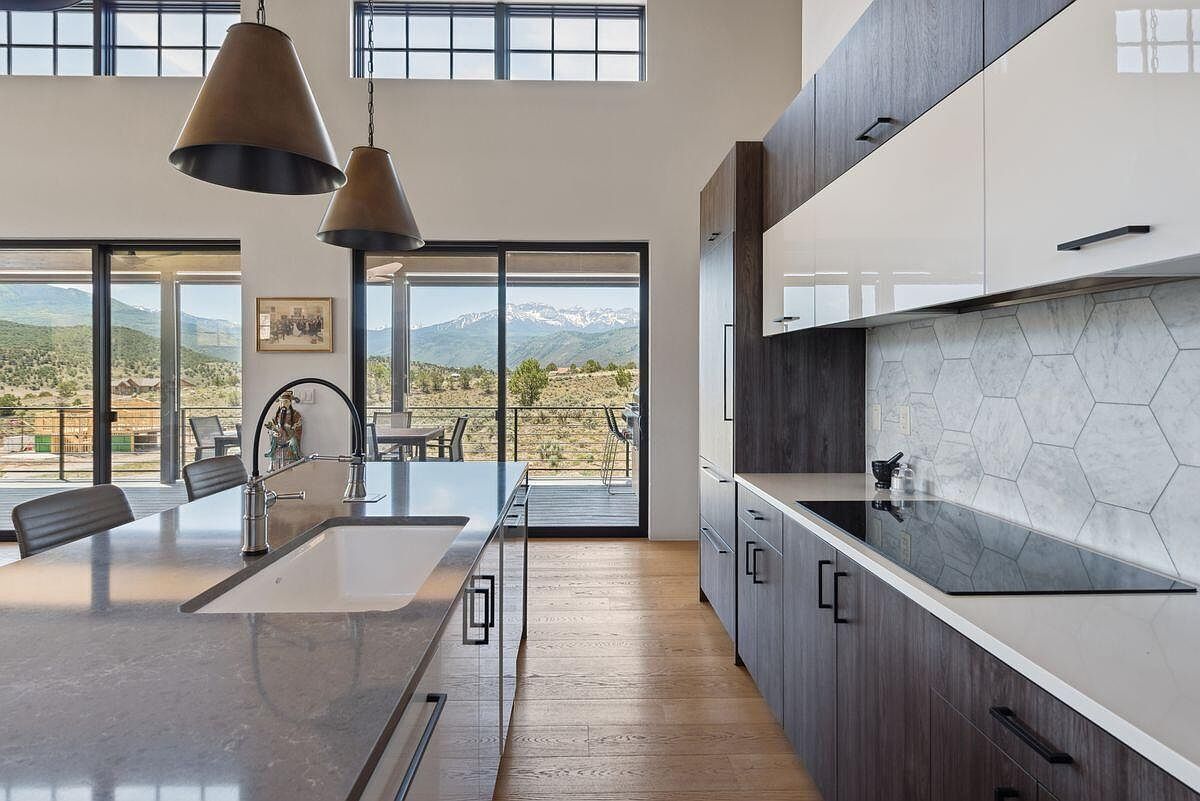 This is a modern kitchen with dark wood and white cabinets, a large island with a sink, and stainless steel appliances. The kitchen features a hexagon tile backsplash and pendant lighting. Large sliding glass doors offer a view of the mountains and outdoor deck.