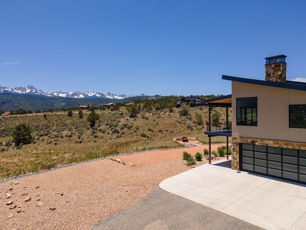 This image showcases the side of a modern home with a focus on its architectural details and surrounding landscape. The house features a stone chimney, a balcony with metal railings, and a garage with a contemporary glass door. The property includes a gravel driveway, native vegetation, and a backdrop of distant mountains, creating a sense of privacy and natural beauty.