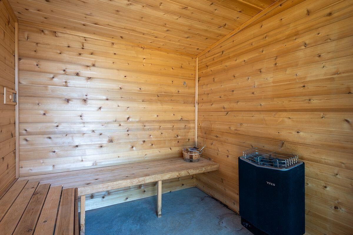 This is an interior shot of a sauna, featuring wooden walls and benches. A black sauna heater stands in the corner, and a small wooden bucket sits on the bench. The room is well-lit, creating a warm and inviting atmosphere, perfect for relaxation and wellness.
