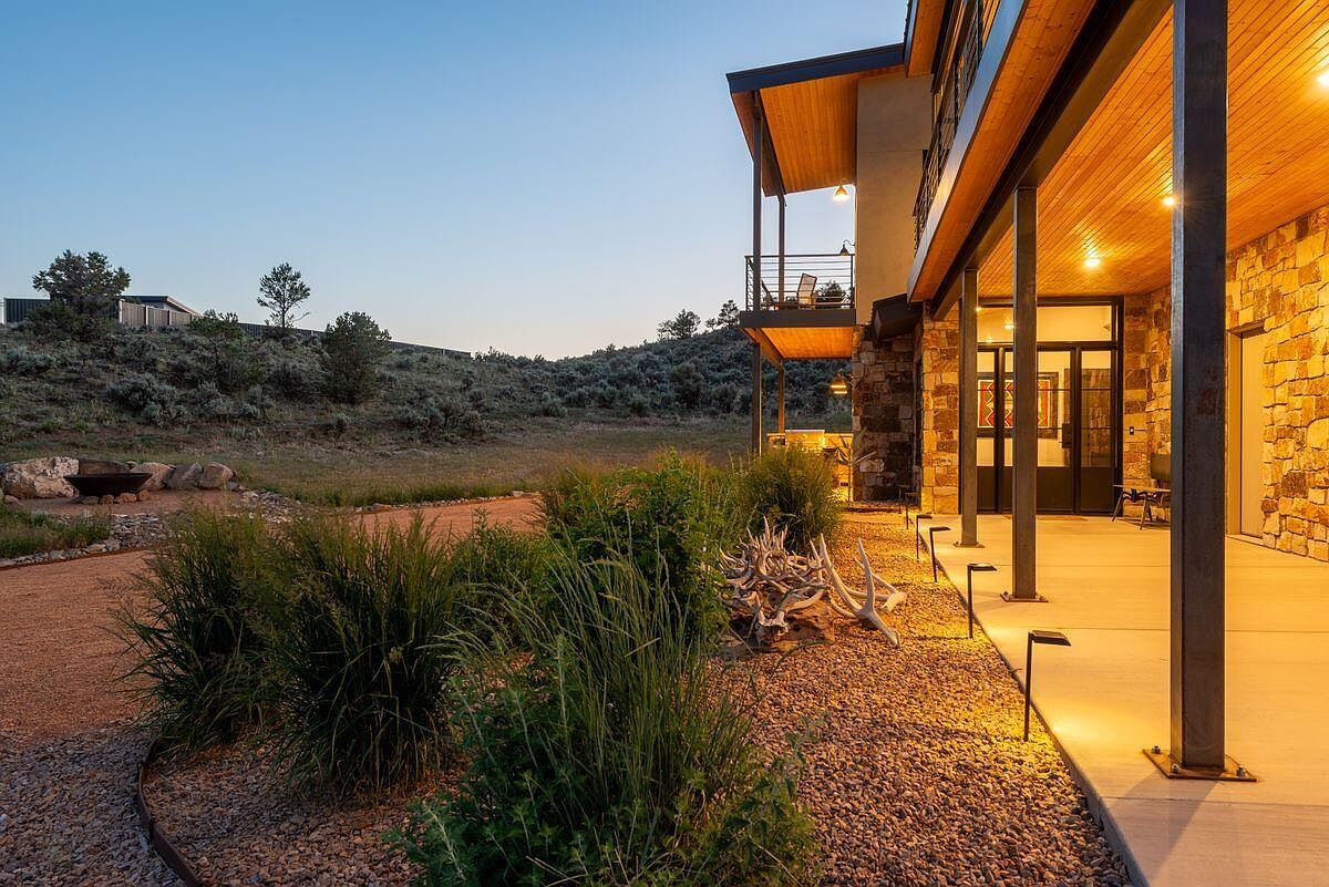 This image showcases the side exterior of a modern home with a stone facade and a covered patio supported by steel columns. The landscape features gravel pathways, ornamental grasses, and natural wood elements, creating a serene and inviting atmosphere. The warm lighting under the patio roof enhances the architectural details and adds to the home's curb appeal.
