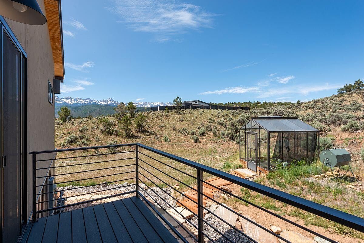 This image showcases a modern deck or balcony with sleek, dark railings and composite decking. A greenhouse and a compost tumbler are visible in the yard, suggesting a focus on gardening and sustainability. In the background, a scenic mountain range adds a touch of natural beauty, enhancing the property's appeal.