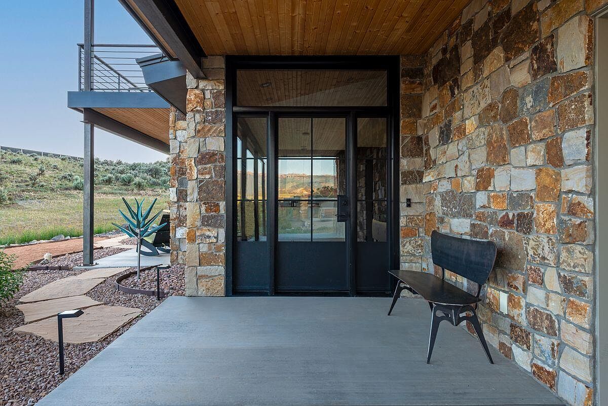 This image showcases a modern home's entryway, featuring a striking stone facade and a sleek black-framed glass door. A concrete porch provides a clean, contemporary feel, complemented by a minimalist metal bench. The exterior is well-lit with landscape lighting, and the surrounding landscape adds to the property's appeal.