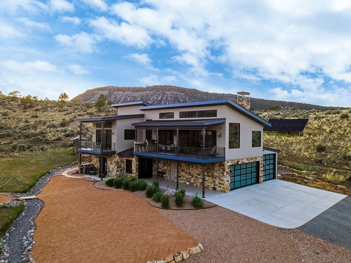 This aerial view showcases a modern two-story home with a combination of stone and stucco exterior. The property features a spacious concrete driveway leading to a garage with glass doors, a landscaped yard with native plants, and balconies on both levels. The house is nestled in a hillside setting with views of the surrounding landscape.