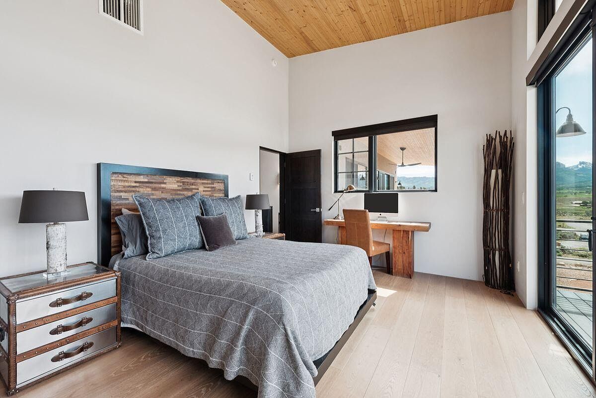 This is a primary bedroom featuring a bed with a wooden headboard and gray bedding. The room has a modern design with a wooden ceiling, light wood floors, and a desk area with a computer. A large window and sliding glass door provide natural light and a view of the outdoors.