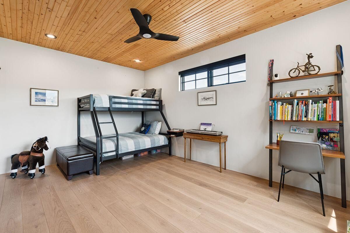 This is a well-lit bedroom featuring a bunk bed with a gray frame and striped bedding. The room also includes a wooden desk with a chair, a bookshelf filled with books and decorative items, and a small ottoman. The wooden ceiling and light wood flooring add warmth to the space, creating a cozy and inviting atmosphere.