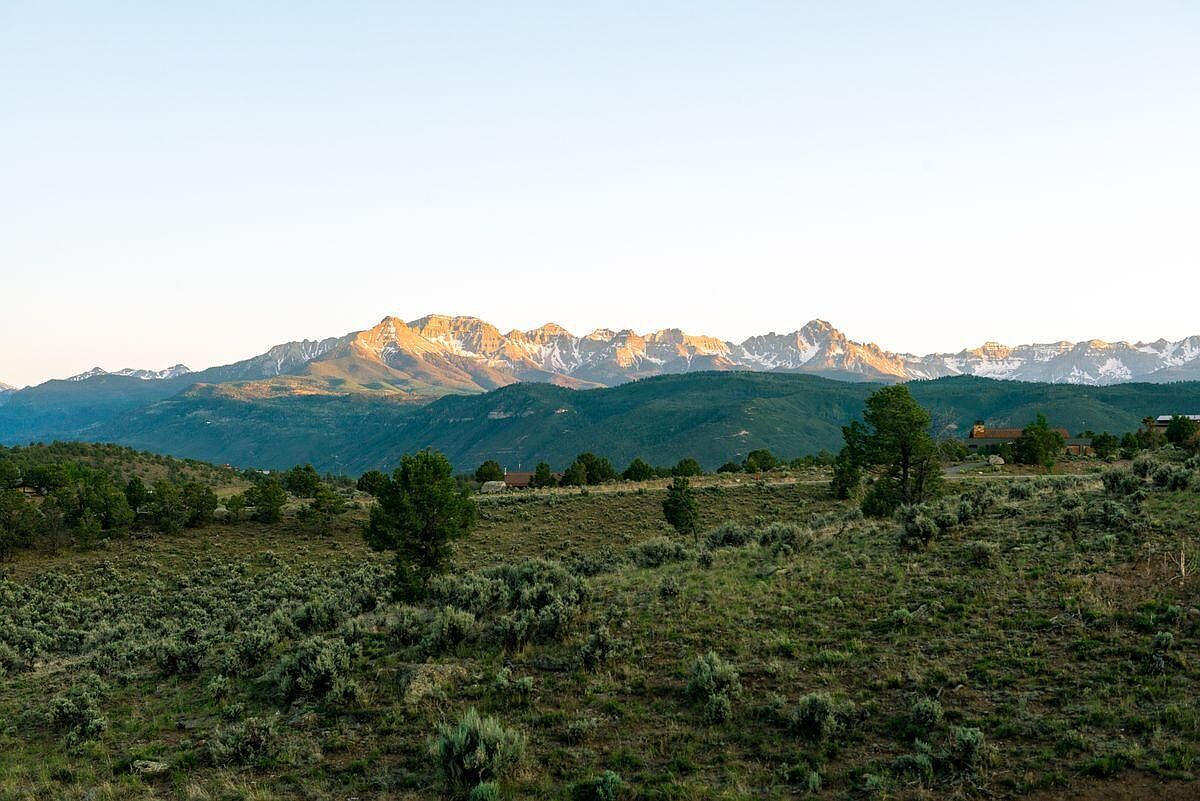 This exterior shot showcases a sprawling yard with native vegetation, set against a backdrop of majestic mountains. The landscape features a mix of grasses and shrubs, creating a natural and serene environment. Several houses are nestled among the trees, suggesting a peaceful and secluded setting.