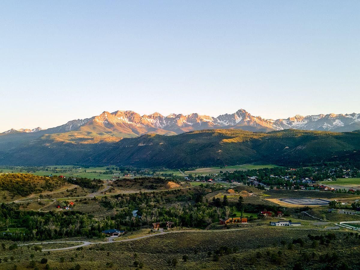 This aerial view showcases a picturesque landscape with a mountain range in the background, partially covered in snow. The foreground features a mix of residential areas and natural terrain, including trees, fields, and winding roads. The overall impression is one of serene beauty and spaciousness, highlighting the property's proximity to both nature and community amenities.
