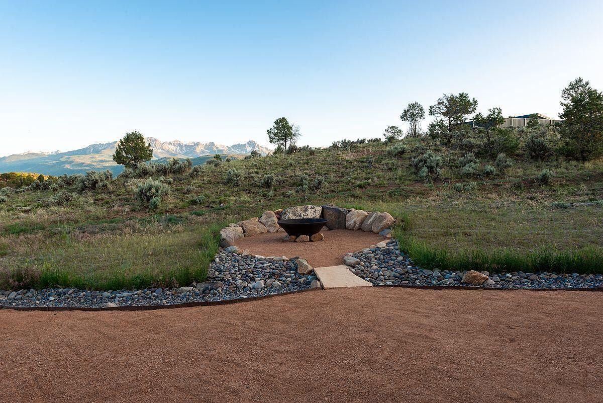 This exterior shot showcases a beautifully landscaped yard featuring a circular fire pit area surrounded by rocks and gravel. A pathway leads to the fire pit, which is set against a backdrop of rolling hills and distant mountains under a clear sky. The scene evokes a sense of tranquility and outdoor living.