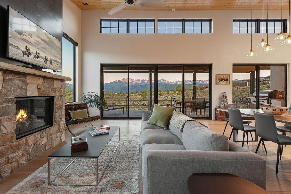 This is an interior shot of a modern living room featuring a stone fireplace with a mounted television, a gray sectional sofa with green pillows, and a glass-topped coffee table on a patterned rug. Large sliding glass doors offer a stunning view of mountains in the distance, and the room is well-lit with natural light and pendant lighting, creating a warm and inviting atmosphere.