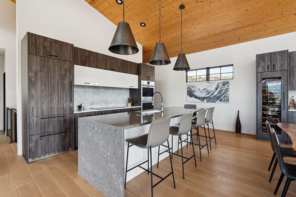 This is a modern kitchen featuring dark wood and white cabinetry, a large gray island with seating, and stainless steel appliances. Three pendant lights hang above the island, and a window provides natural light and a view of the mountains. The kitchen has a clean and contemporary style with wood floors and a wood ceiling.