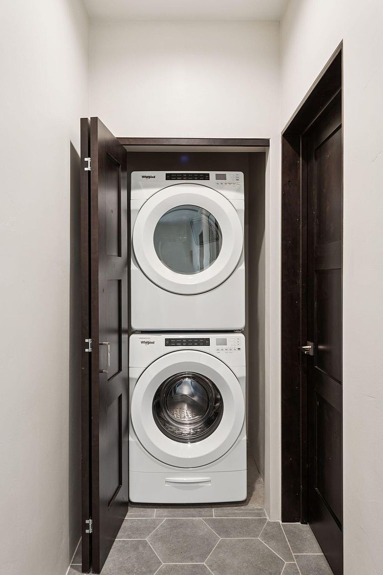 This is an interior shot of a laundry room featuring a stacked Whirlpool washer and dryer unit. The appliances are white and neatly fitted into a dark wood cabinet. The floor is tiled with gray hexagonal tiles, and a partially open dark wood door is visible to the left, adding depth to the space.
